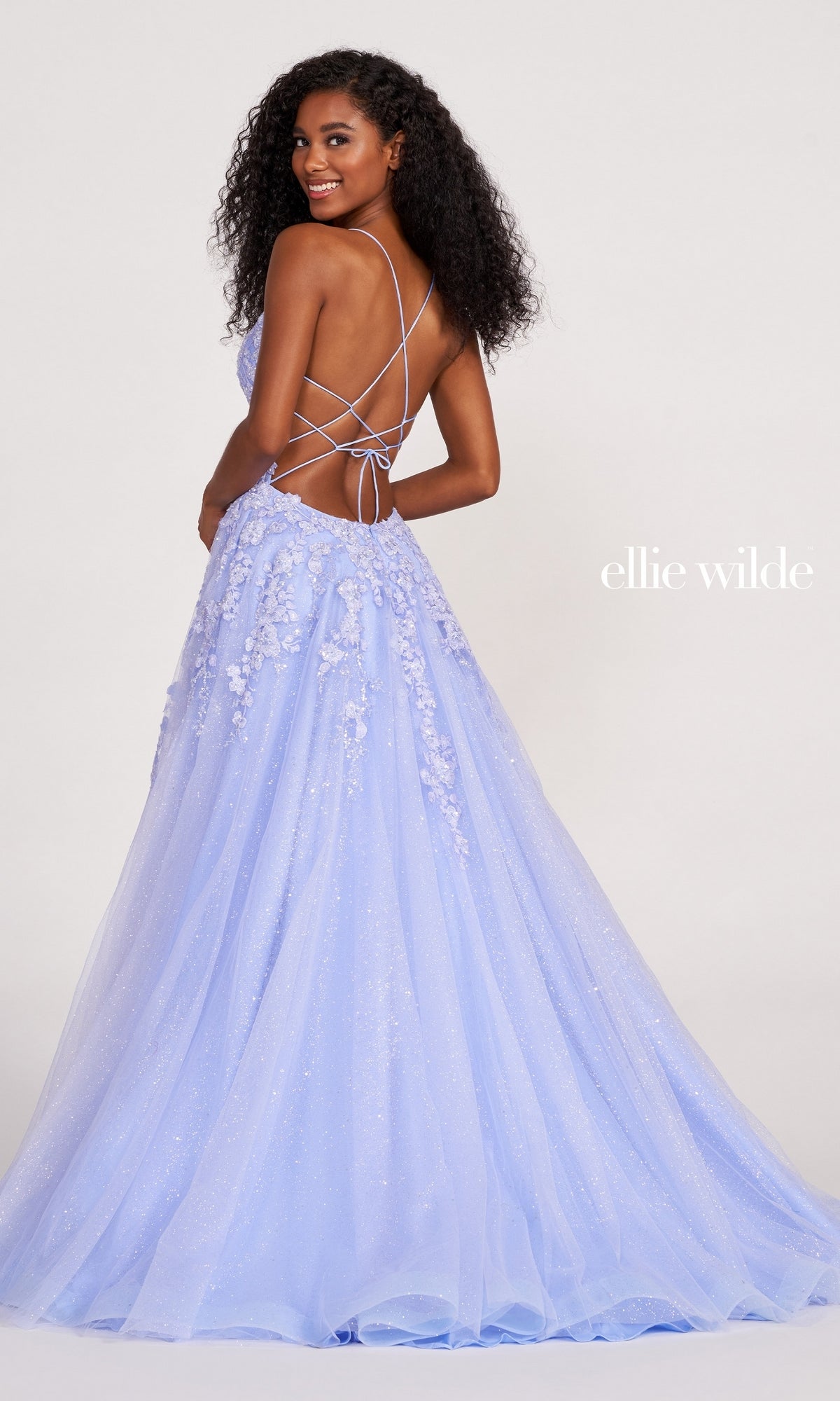 A woman in the Ellie Wilde Backless Designer Prom Dress EW34053, light purple with floral appliqué and a lace-up back, stands facing away, looking over her shoulder and smiling.